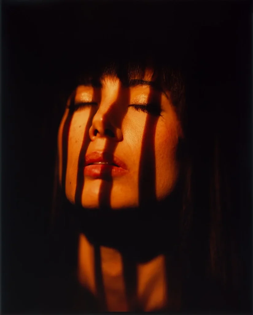 Close-up 8x10 film portrait of a woman with her eyes closed, warm low sunlight striped across her face by window blinds against a dark background