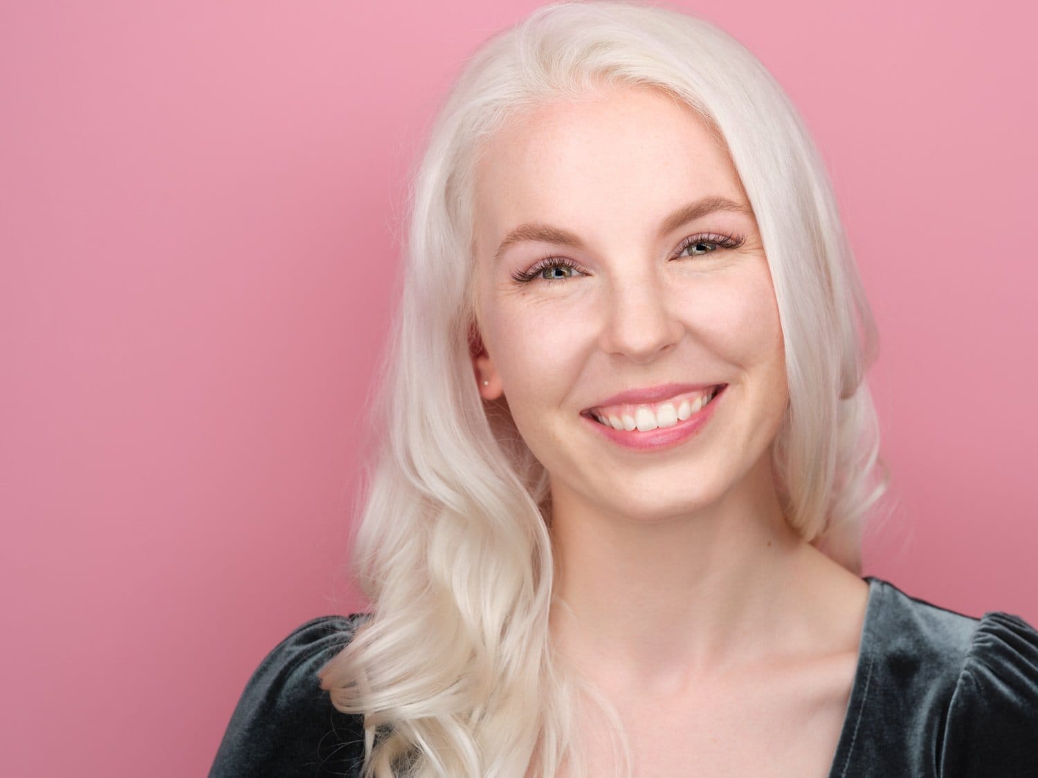 A woman with long, wavy blonde hair smiling against a pink background.