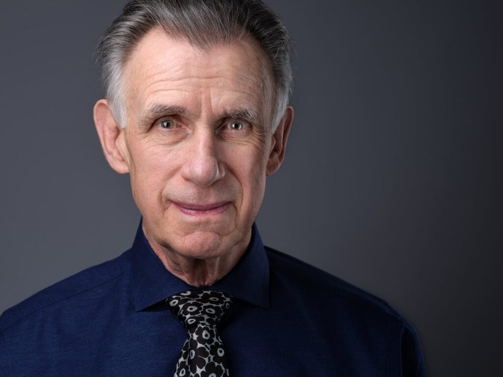Business headshot of an older man in a dark navy shirt and patterned tie against a grey studio background