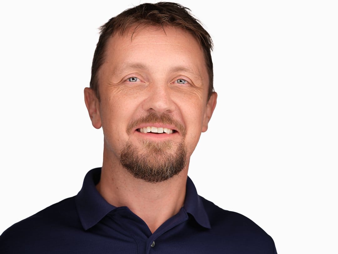 Headshot of a man with light skin, short brown hair, blue eyes, and a goatee, smiling, wearing a navy blue collared shirt, against a white background.