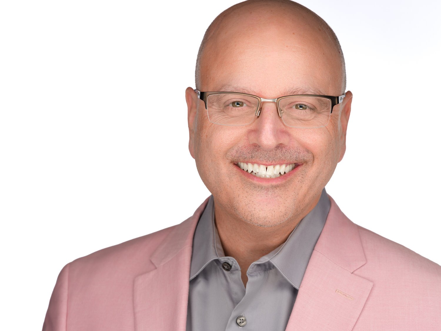 A smiling man with glasses, wearing a gray shirt and a pink blazer, against a white background.