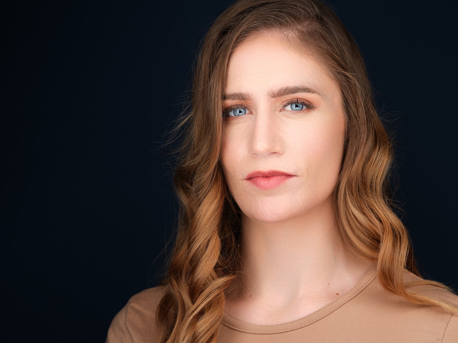 Close-up portrait of a woman with wavy light brown hair, blue eyes, and fair skin, wearing a beige top, against a dark background.