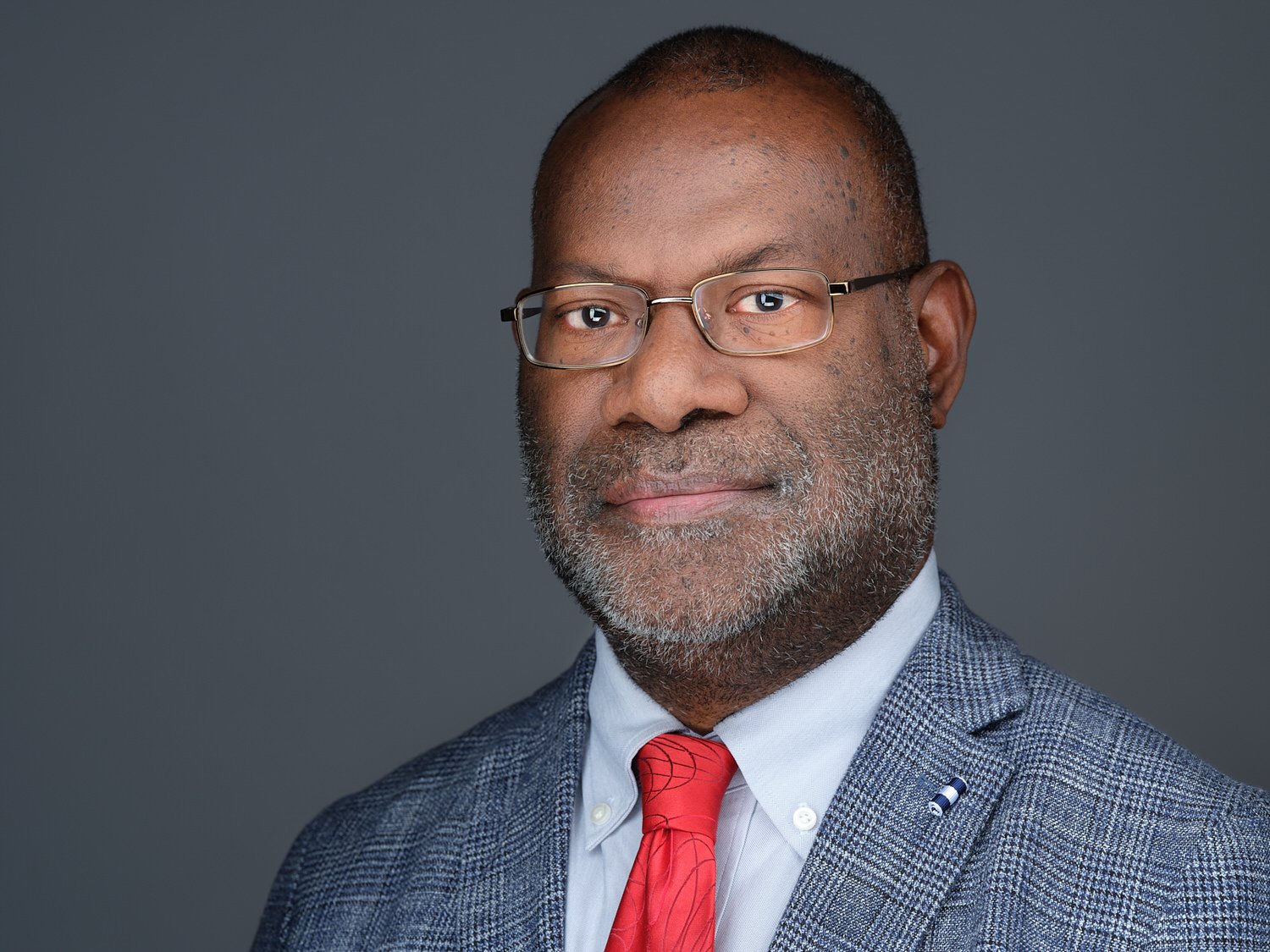 Close-up portrait of a middle-aged African American man with gray facial hair, wearing glasses, a gray plaid suit, a white shirt, and a red tie, against a dark gray background.