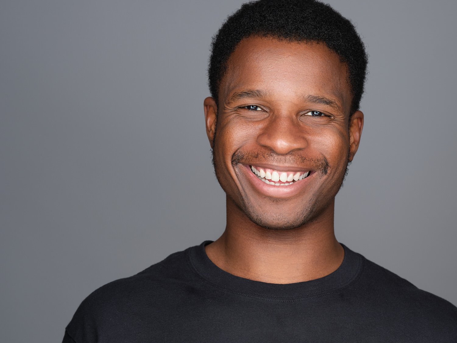 Smiling Black man with short curly hair wearing a black t-shirt against a gray background.