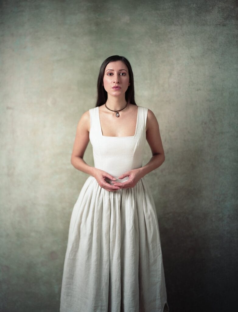 Full-length 8x10 film portrait of a woman in a cream linen dress standing against a mottled green painted backdrop, hands clasped in front of her