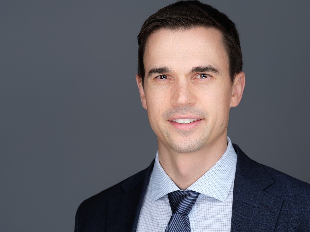 Professional headshot of a young man in a suit and tie, smiling against a gray background.
