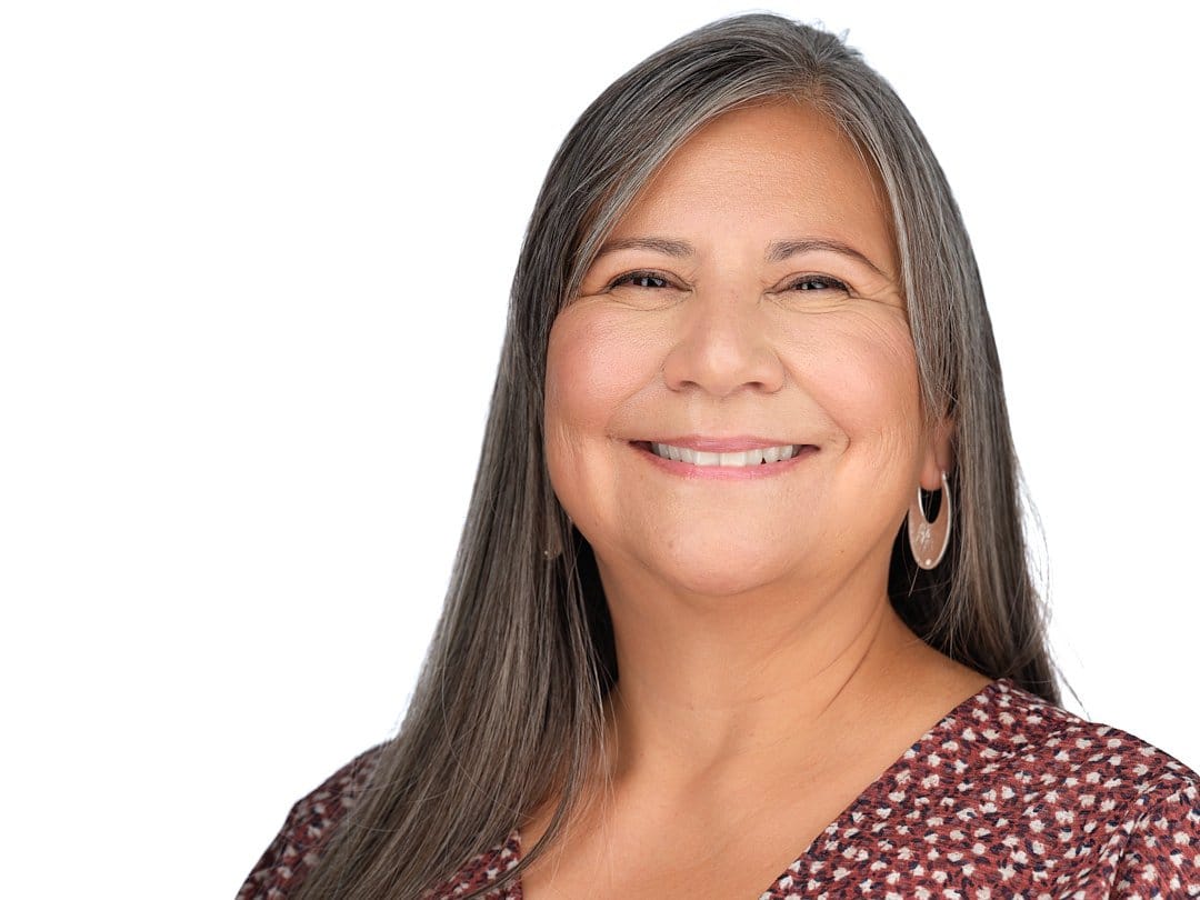 Portrait of a smiling middle-aged woman with long gray hair, wearing hoop earrings and a patterned blouse, against a plain white background.
