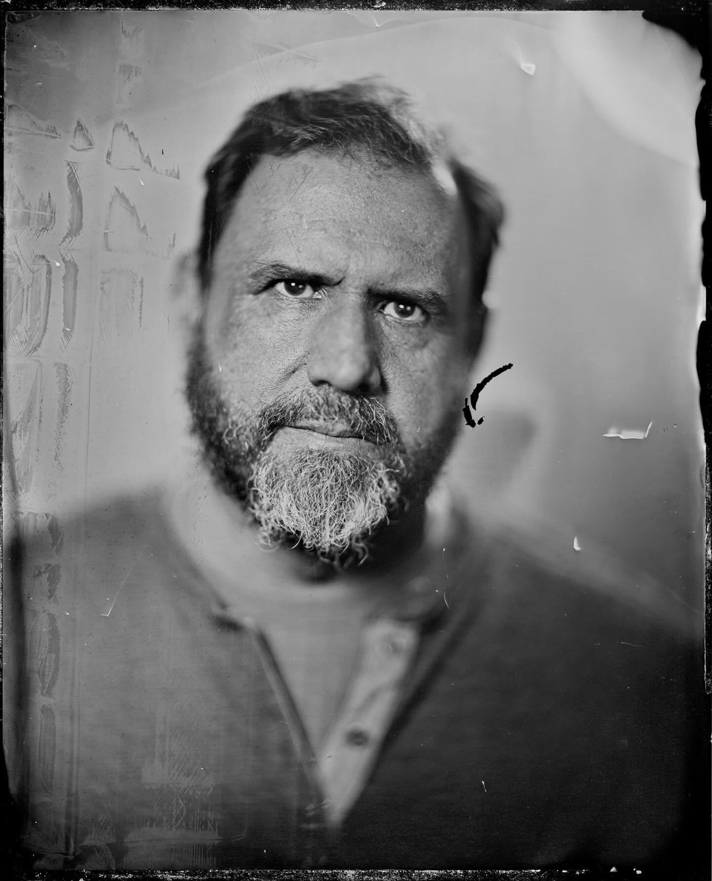 Black and white close-up photograph of an older man with a beard and short hair, looking directly at the camera with a serious expression.