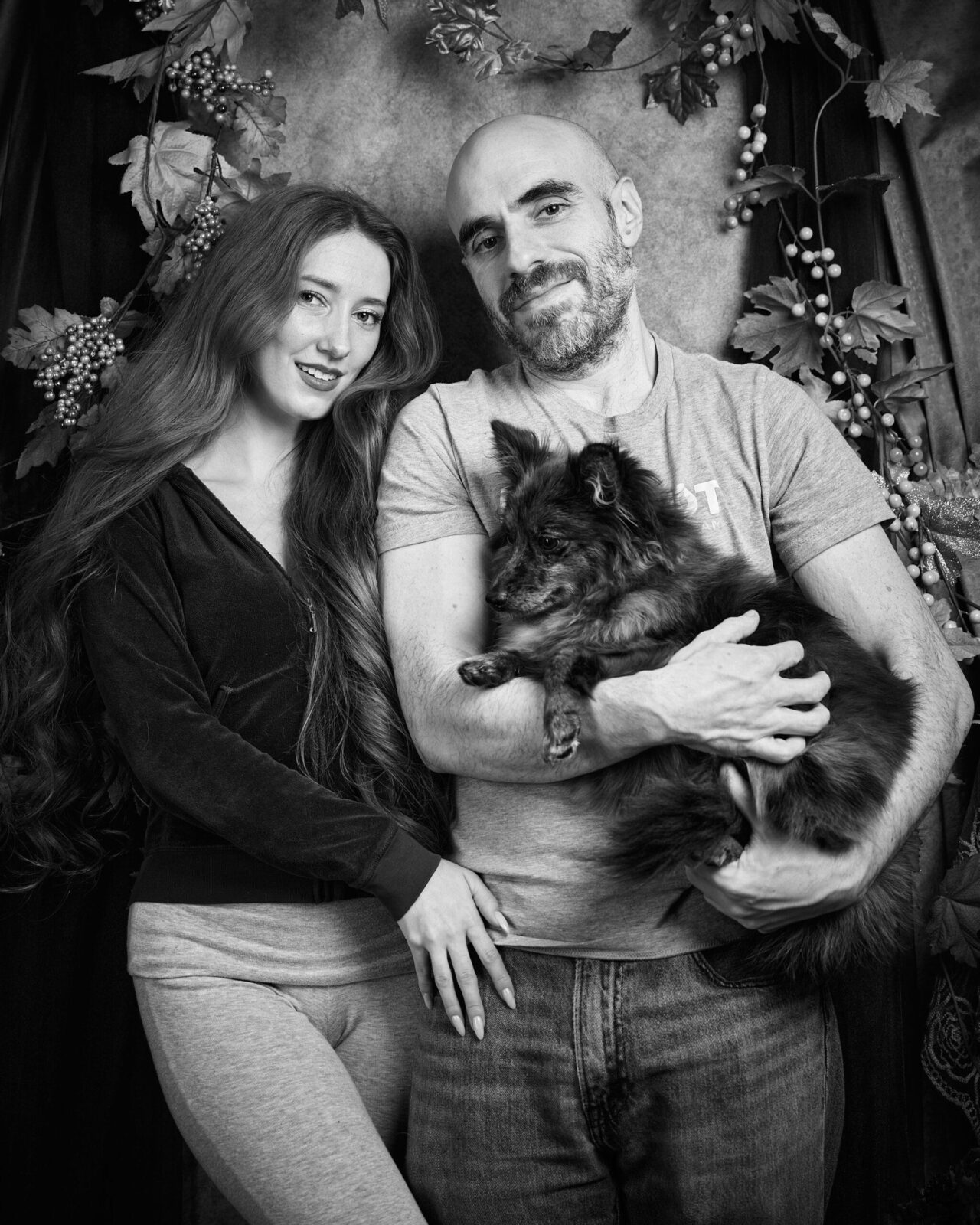 A black and white photo of a smiling woman with long wavy hair, a man with a bald head and beard holding a small fluffy dog, standing close together in front of a decorative backdrop with foliage and berries.