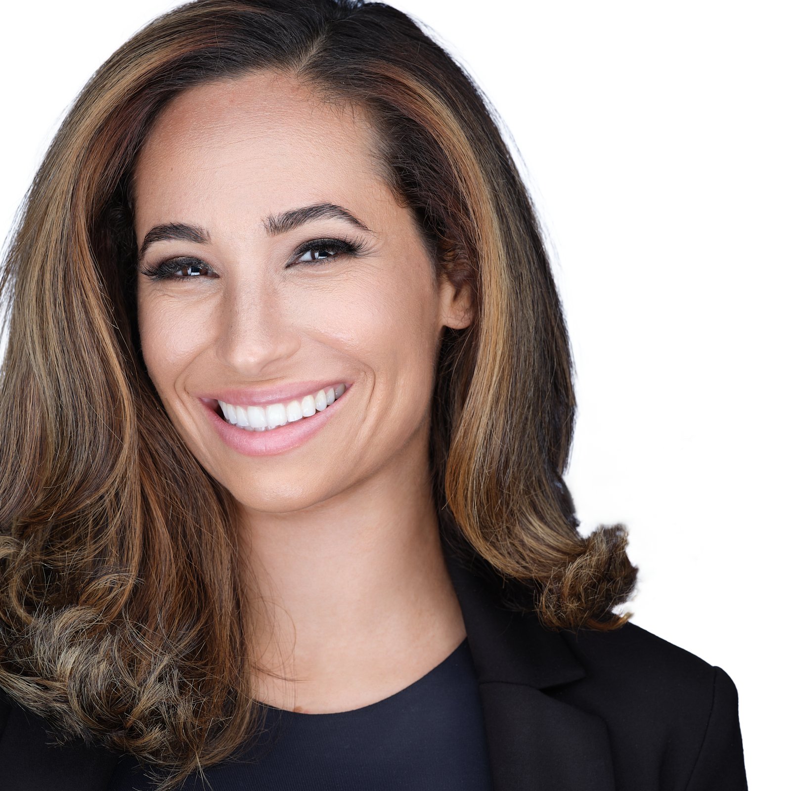 Close-up of a smiling woman with wavy brown hair, wearing a black blazer over a dark top, against a white background.