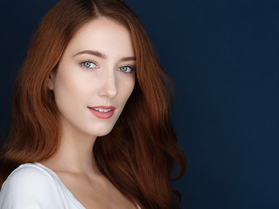 Close-up portrait of a woman with long red hair, blue eyes, and fair skin, smiling softly, wearing a white top, against a dark background.