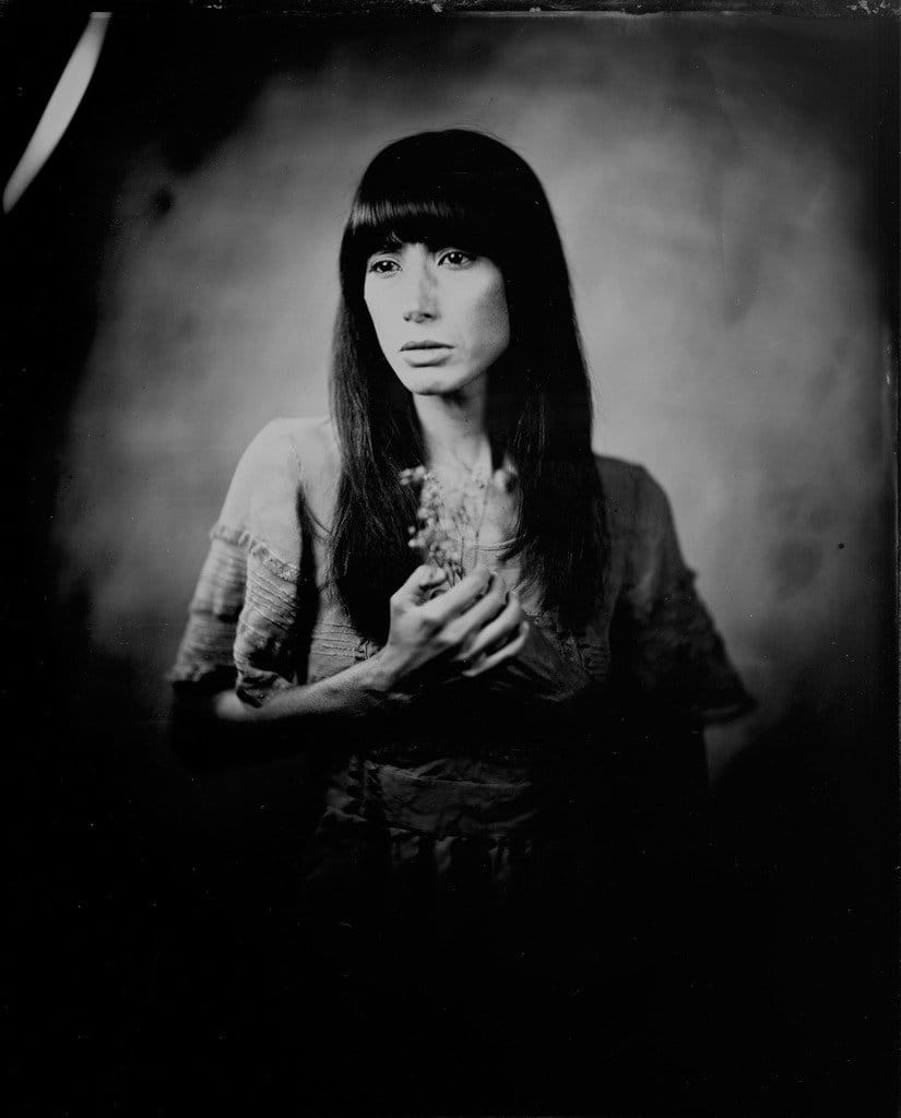 Black and white portrait of a woman with long, straight dark hair and bangs, holding a small plant or flower in her hands, looking slightly to the right.